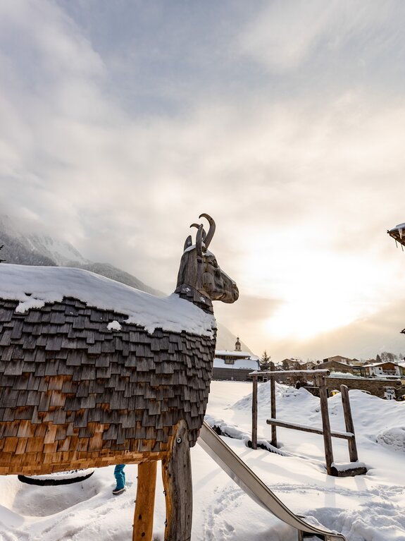 Holzstatue eines Steinbocks mit Schnee auf dem Dach vor verschneiter Berglandschaft