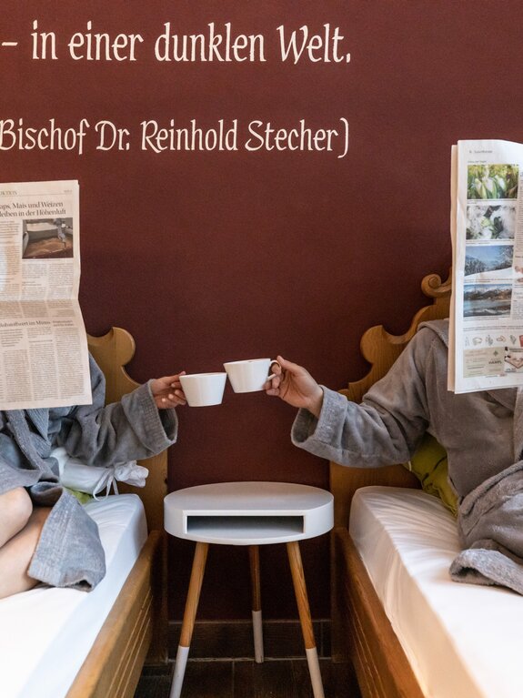 Zwei Personen im Bademantel mit Zeitung und Kaffeetassen im Bett