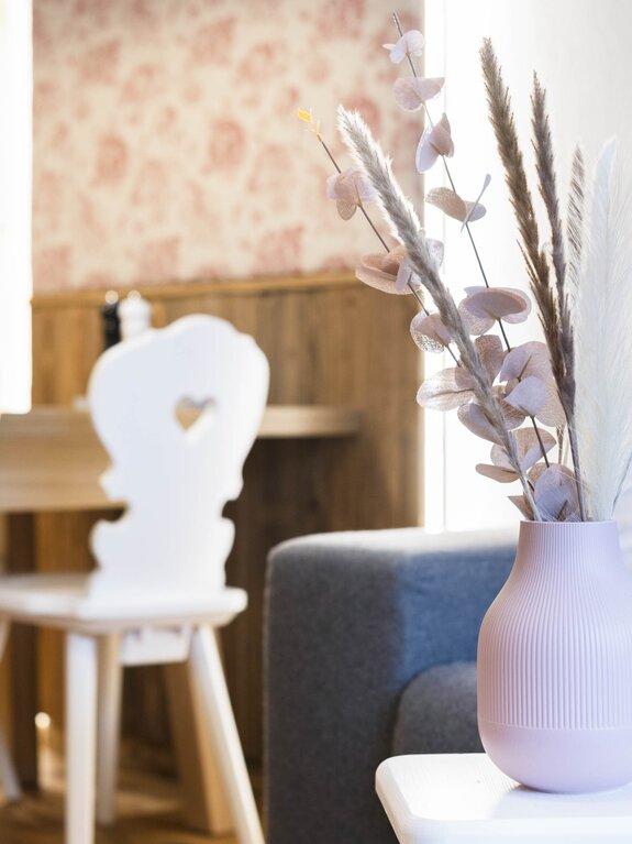 Rosa Vase mit Trockenblumen auf Tisch, weiße Stühle mit Herz, graues Sofa im Hintergrund