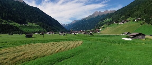 Tal mit Dorf zwischen grünen Wiesen und bewaldeten Bergen unter blauem Himmel